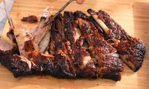 Costillas a la barbacoa jugosas y tiernas, cortadas en una tabla de madera. La carne se desprende del hueso, cubierta de salsa.
