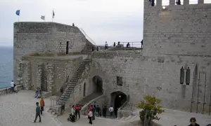 El castillo de Peñíscola se alza imponente sobre el mar. Turistas pasean por sus patios de piedra, admirando la arquitectura histórica.
