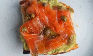 Tostada con aguacate y generosas lonchas de salmón marinado, aderezada con alcaparras y pimienta negra.