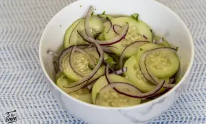 Deliciosa ensalada de pepino con cebolla morada en un tazón blanco sobre un mantel azul y blanco.