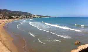 La **playa romana alcossebre** se extiende con arena dorada y aguas turquesas bajo un cielo azul. Olas suaves rompen en la orilla, mientras la costa se dibuja con casas y montañas al fondo.