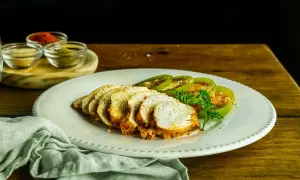 Pechugas de pollo al horno, jugosas y doradas, servidas con rodajas de tomate verde y perejil fresco. Especias en pequeños cuencos al fondo.