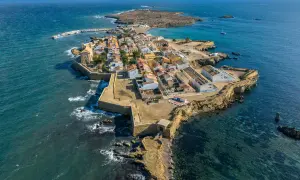 Vista aérea de una pintoresca cala del sur, con casas blancas y tejados naranjas, rodeada por el mar azul y una fortaleza.