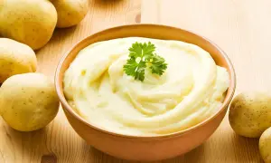 Un cremoso pure de patatas, decorado con perejil, junto a patatas frescas sobre una mesa de madera.