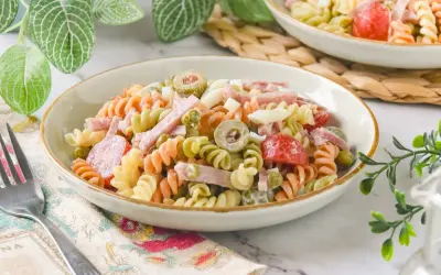 Un plato de ensalada de pasta tricolor con jamón, huevo duro, aceitunas y guisantes.