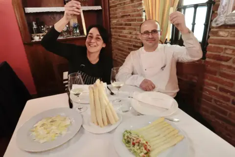 Pareja sonríe mostrando espárragos blancos, uno de ellos en un plato con ensalada y el otro en un manojo atado.