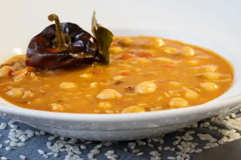 Un plato humeante de garbanzos con arroz, decorado con un pimiento seco y una hoja de laurel.