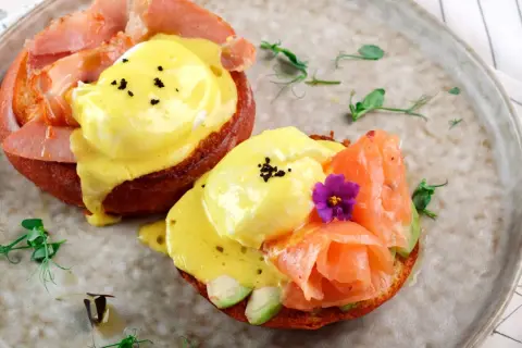 Dos deliciosos huevos benedictinos, uno con jamón y otro con salmón ahumado y aguacate, servidos sobre pan tostado con salsa holandesa.