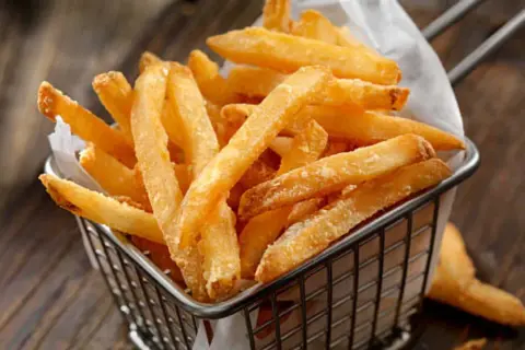 Cesta metálica llena de crujientes patatas fritas al horno, servidas sobre papel blanco.