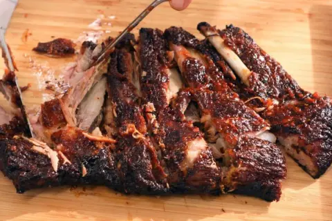 Costillas a la barbacoa jugosas y tiernas, cortadas en una tabla de madera. La carne se desprende del hueso, cubierta de salsa.