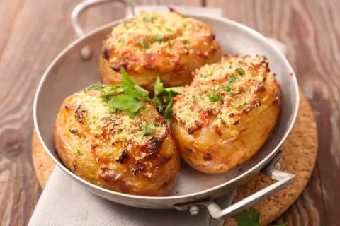 Tres patatas rellenas doradas y crujientes, cubiertas con queso rallado y perejil fresco, servidas en una sartén metálica.