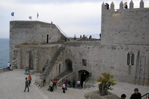 El castillo de Peñíscola se alza imponente sobre el mar. Turistas pasean por sus patios de piedra, admirando la arquitectura histórica.