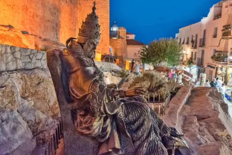 Estatua del Papa Luna sentado, con el atardecer iluminando el fondo y gente paseando. Se rumorea cómo murió el Papa Luna.
