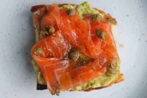 Tostada con aguacate y generosas lonchas de salmón marinado, aderezada con alcaparras y pimienta negra.