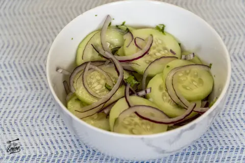 Deliciosa ensalada de pepino con cebolla morada en un tazón blanco sobre un mantel azul y blanco.