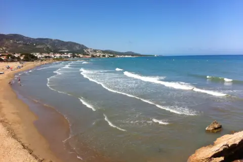 La **playa romana alcossebre** se extiende con arena dorada y aguas turquesas bajo un cielo azul. Olas suaves rompen en la orilla, mientras la costa se dibuja con casas y montañas al fondo.