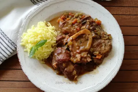 Un plato de osso buco con arroz amarillo y una hoja de albahaca. La carne está cubierta de una salsa con verduras.
