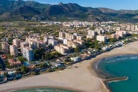 Vista aérea del casco antiguo de Benicàssim pueblo, con edificios residenciales, playas de arena y el mar Mediterráneo. Montañas verdes al fondo.