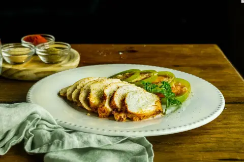 Pechugas de pollo al horno, jugosas y doradas, servidas con rodajas de tomate verde y perejil fresco. Especias en pequeños cuencos al fondo.