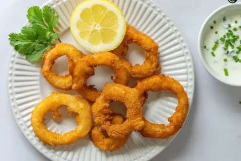 Crujientes aros de cebolla dorados, servidos con una rodaja de limón y un dip cremoso. ¡Un aperitivo irresistible!