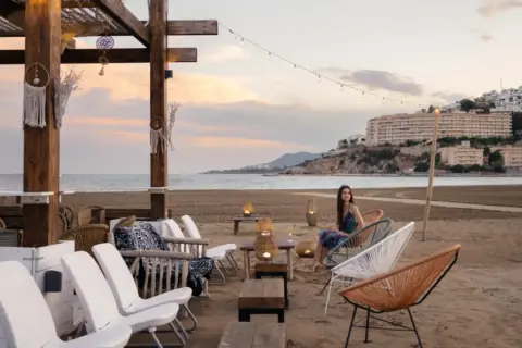 Una mujer disfruta de la tarde en la playa sur Peñíscola, sentada en una silla de diseño frente al mar.
