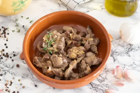 Mollejas de pollo guisadas con ajo y tomillo en cazuela de barro, acompañadas de granos de pimienta y una cabeza de ajo.