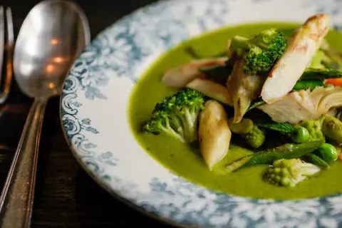 Deliciosa menestra de verduras en un plato con borde azul y blanco. Brócoli, espárragos y guisantes flotan en una cremosa salsa verde.