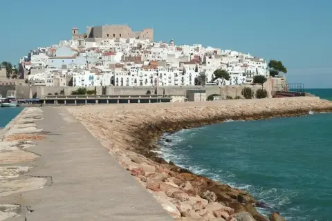 Pueblos costa Azahar: un castillo y casas blancas se alzan sobre una colina, con el mar turquesa y un rompeolas de rocas.