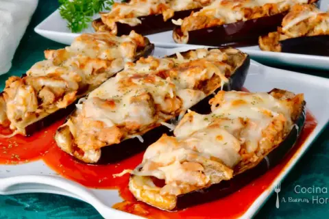 Berenjenas rellenas de atún gratinadas con queso, servidas sobre una base de salsa roja.