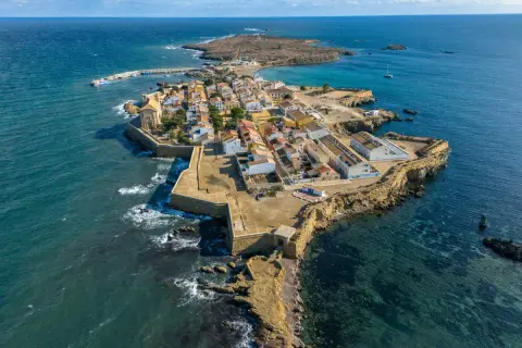 Vista aérea de una pintoresca cala del sur, con casas blancas y tejados naranjas, rodeada por el mar azul y una fortaleza.