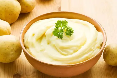 Un cremoso pure de patatas, decorado con perejil, junto a patatas frescas sobre una mesa de madera.