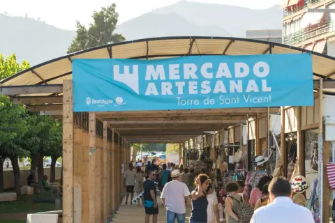 Gente pasea por un mercadillo en Benicàssim, bajo un cartel azul que anuncia "Mercado Artesanal Torre de Sant Vicent".