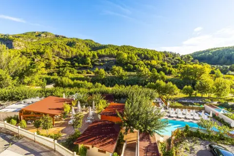 Un día soleado en la rosaleda de Mijares, con un complejo turístico y piscina rodeado de exuberante vegetación y montañas verdes.