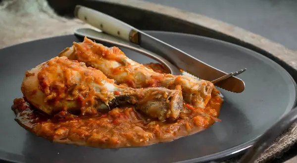 Dos muslos de pollo al chilindrón, servidos en un plato oscuro con cubiertos.