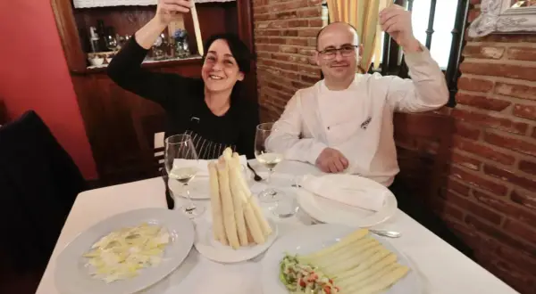 Pareja sonríe mostrando espárragos blancos, uno de ellos en un plato con ensalada y el otro en un manojo atado.