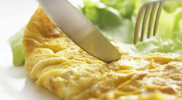 Un cuchillo corta una dorada tortilla francesa junto a hojas de lechuga fresca.