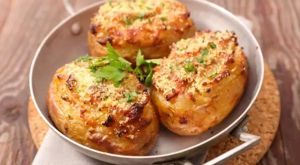 Tres patatas rellenas doradas y crujientes, cubiertas con queso rallado y perejil fresco, servidas en una sartén metálica.