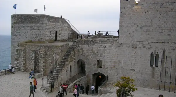 El castillo de Peñíscola se alza imponente sobre el mar. Turistas pasean por sus patios de piedra, admirando la arquitectura histórica.