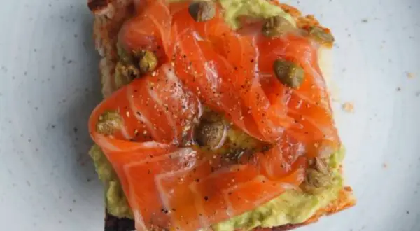 Tostada con aguacate y generosas lonchas de salmón marinado, aderezada con alcaparras y pimienta negra.