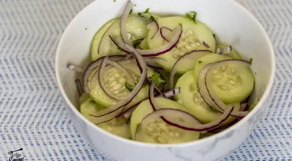 Deliciosa ensalada de pepino con cebolla morada en un tazón blanco sobre un mantel azul y blanco.