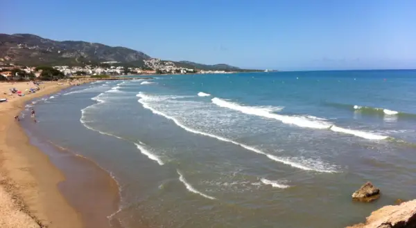 La **playa romana alcossebre** se extiende con arena dorada y aguas turquesas bajo un cielo azul. Olas suaves rompen en la orilla, mientras la costa se dibuja con casas y montañas al fondo.