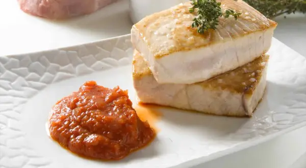 Dos trozos de atún a la plancha apilados, con una guarnición de salsa de tomate y una ramita de tomillo.