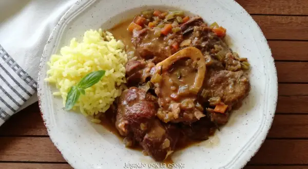 Un plato de osso buco con arroz amarillo y una hoja de albahaca. La carne está cubierta de una salsa con verduras.