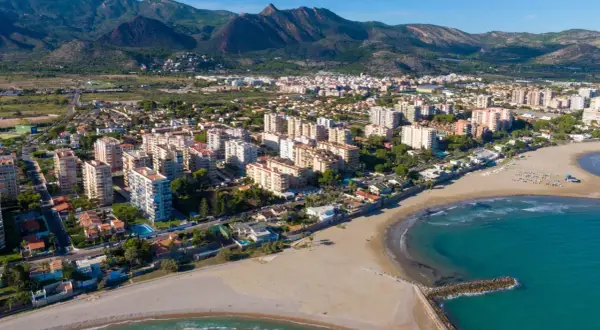 Vista aérea del casco antiguo de Benicàssim pueblo, con edificios residenciales, playas de arena y el mar Mediterráneo. Montañas verdes al fondo.