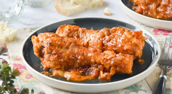 Muslos de pollo en salsa, una receta de la abuela que evoca recuerdos de hogar. Servidos en un plato oscuro, con pan y un tenedor.