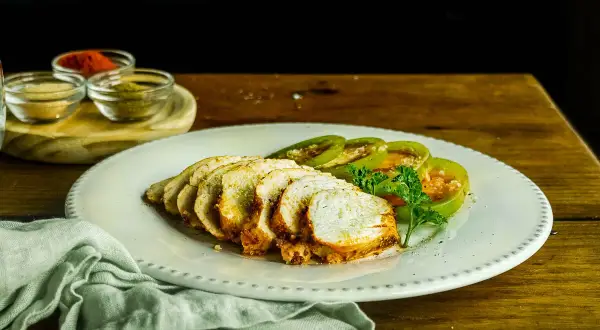 Pechugas de pollo al horno, jugosas y doradas, servidas con rodajas de tomate verde y perejil fresco. Especias en pequeños cuencos al fondo.