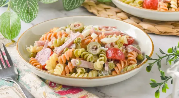 Un plato de ensalada de pasta tricolor con jamón, huevo duro, aceitunas y guisantes.