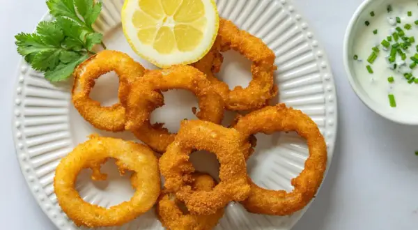 Crujientes aros de cebolla dorados, servidos con una rodaja de limón y un dip cremoso. ¡Un aperitivo irresistible!