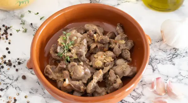 Mollejas de pollo guisadas con ajo y tomillo en cazuela de barro, acompañadas de granos de pimienta y una cabeza de ajo.