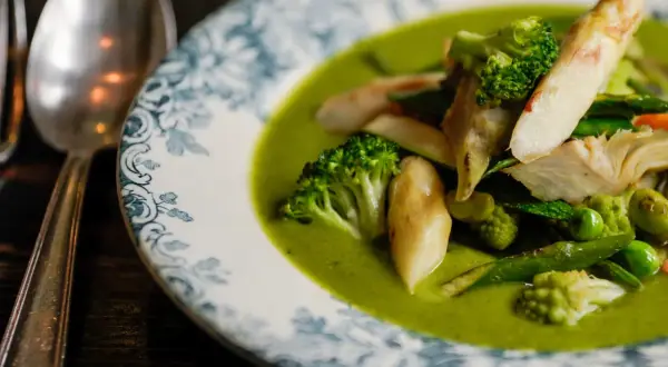 Deliciosa menestra de verduras en un plato con borde azul y blanco. Brócoli, espárragos y guisantes flotan en una cremosa salsa verde.